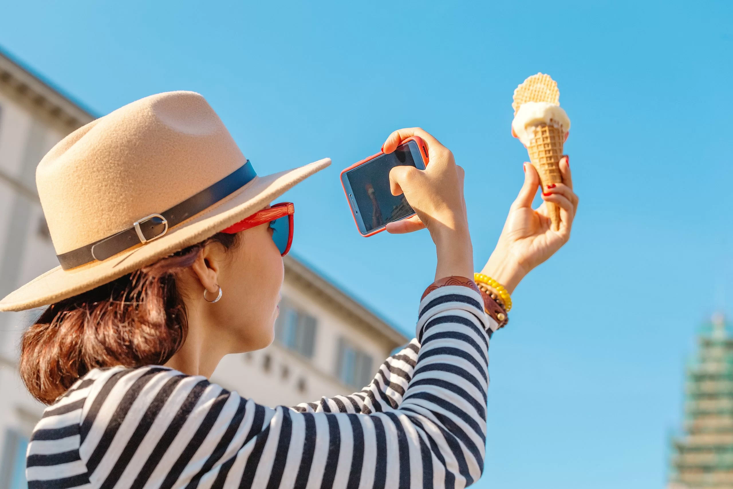 Woman taking a picture with her cell phone of an ice cream cone in front of a blue sky